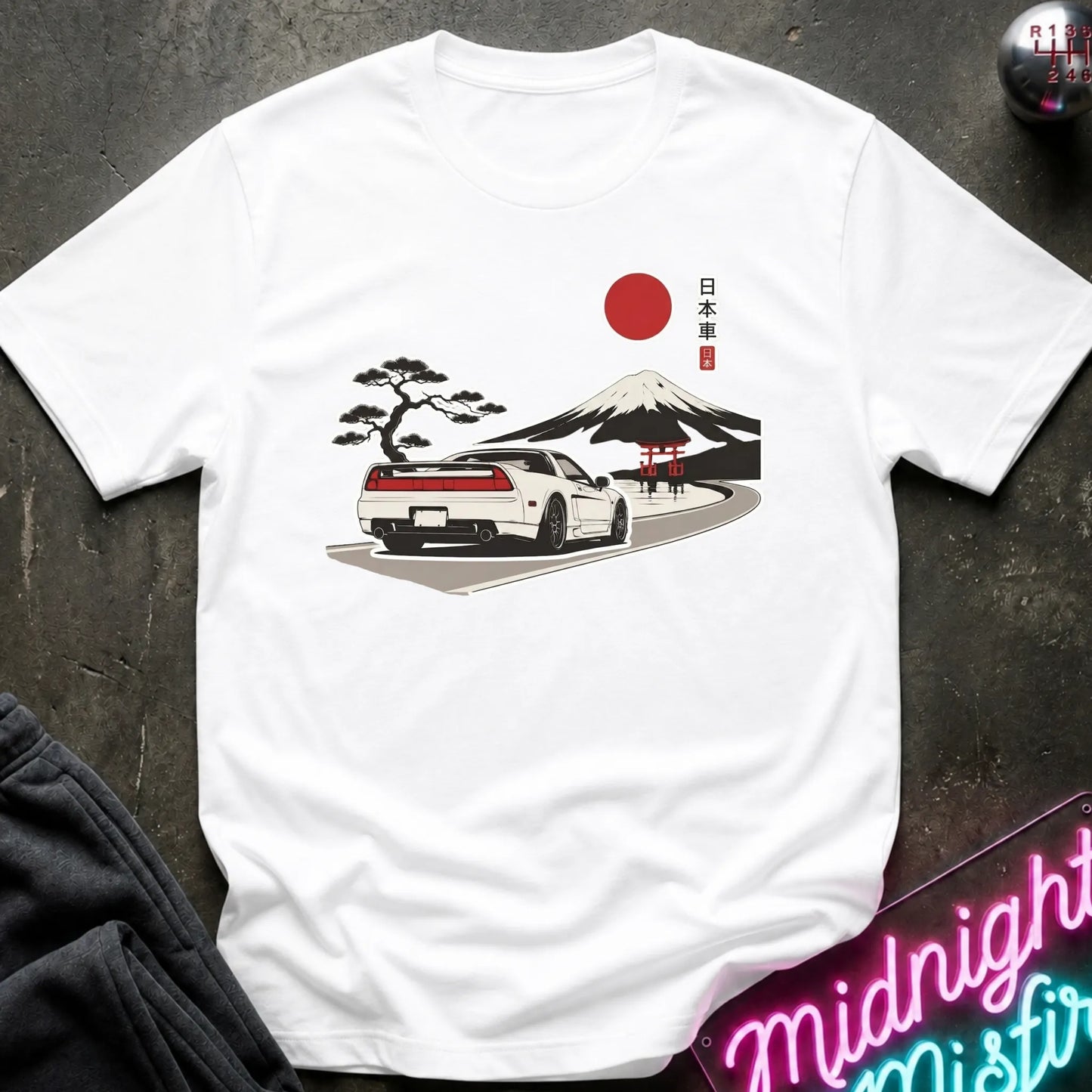 A white short-sleeve t-shirt laid flat on a dark textured surface, featuring a graphic print of a white sports car on a road with a red sun, Mount Fuji, a black pine tree, and a red torii gate, alongside Japanese text.