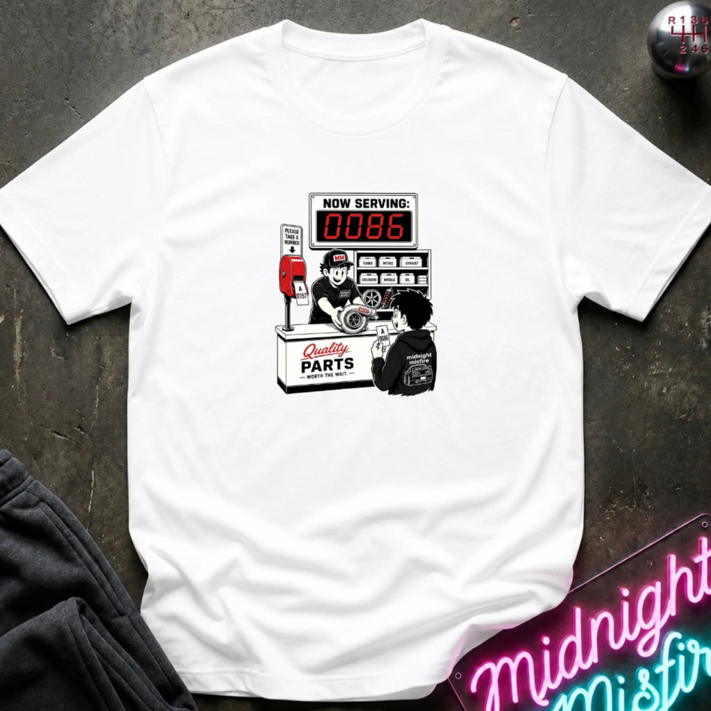 A white  short-sleeve t-shirt with a retro-style graphic of a garage attendant at a 'Quality Parts' counter, displayed flat on a dark textured surface with a neon sign and sweatpants partially visible.