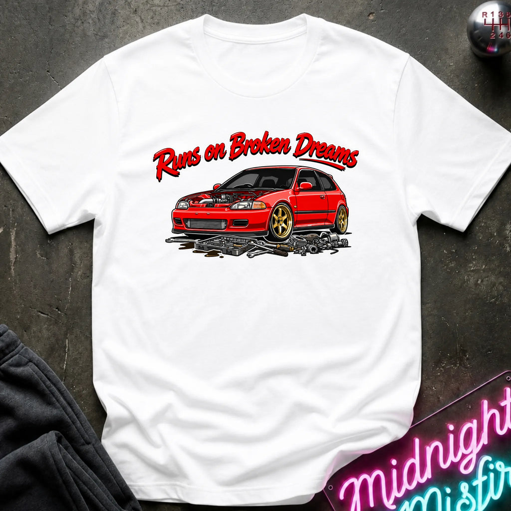 white graphic t-shirt laid flat on a dark textured surface. The shirt features a red sports car with gold wheels and the text 'Runs on Broken Dreams' in red and white.