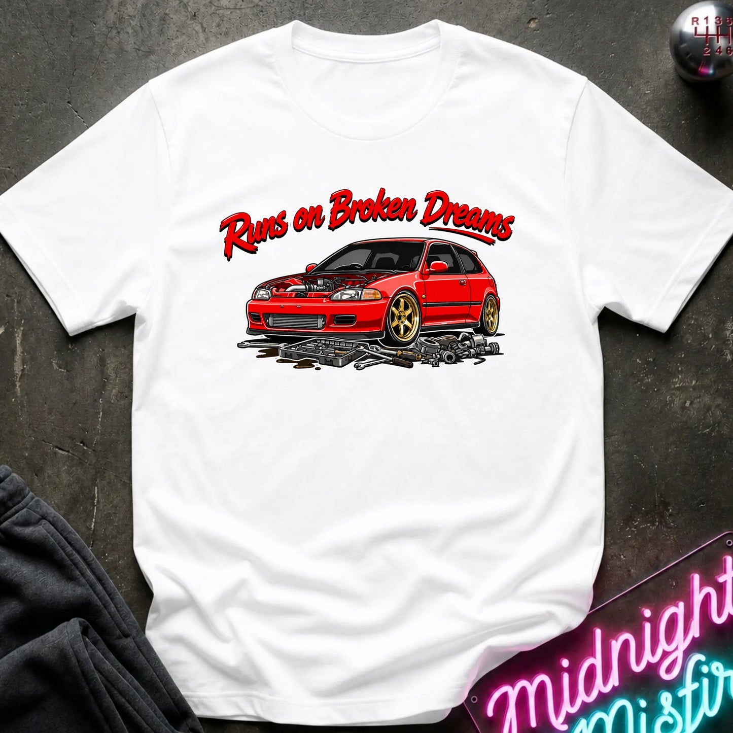 white graphic t-shirt laid flat on a dark textured surface. The shirt features a red sports car with gold wheels and the text 'Runs on Broken Dreams' in red and white.