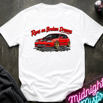 white graphic t-shirt laid flat on a dark textured surface. The shirt features a red sports car with gold wheels and the text 'Runs on Broken Dreams' in red and white.