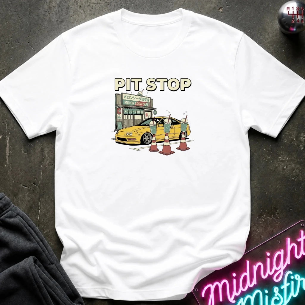 A white short-sleeve t-shirt laid flat on a dark textured surface, featuring a graphic print of a yellow sports car at a pit stop with the text 'PIT STOP' above it. A neon sign and dark clothing are partially visible as props.