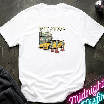 A white short-sleeve t-shirt laid flat on a dark textured surface, featuring a graphic print of a yellow sports car at a pit stop with the text 'PIT STOP' above it. A neon sign and dark clothing are partially visible as props.