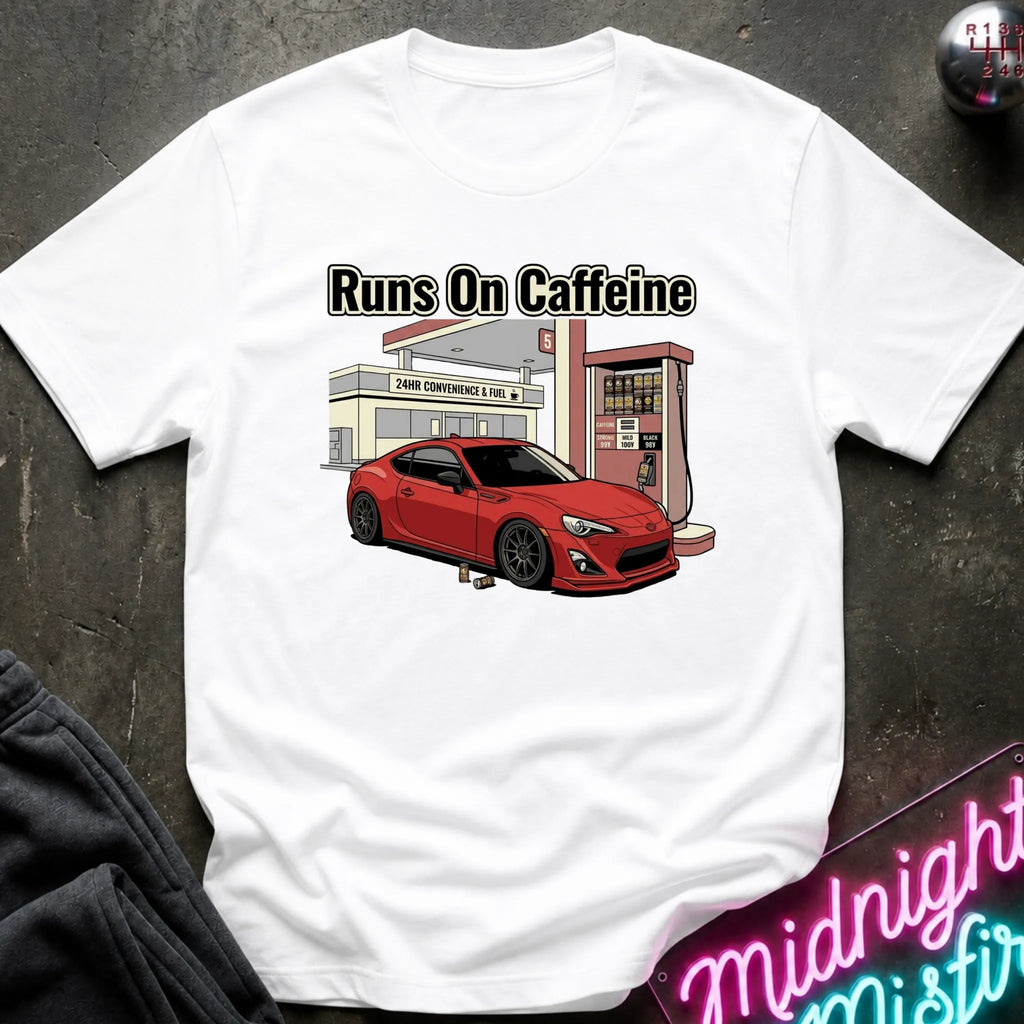 A white short-sleeve graphic T-shirt laid flat on a dark textured surface. The shirt features a printed illustration of a red sports car parked in front of a convenience store and gas station, with the text 'Runs On Caffeine' above the graphic.