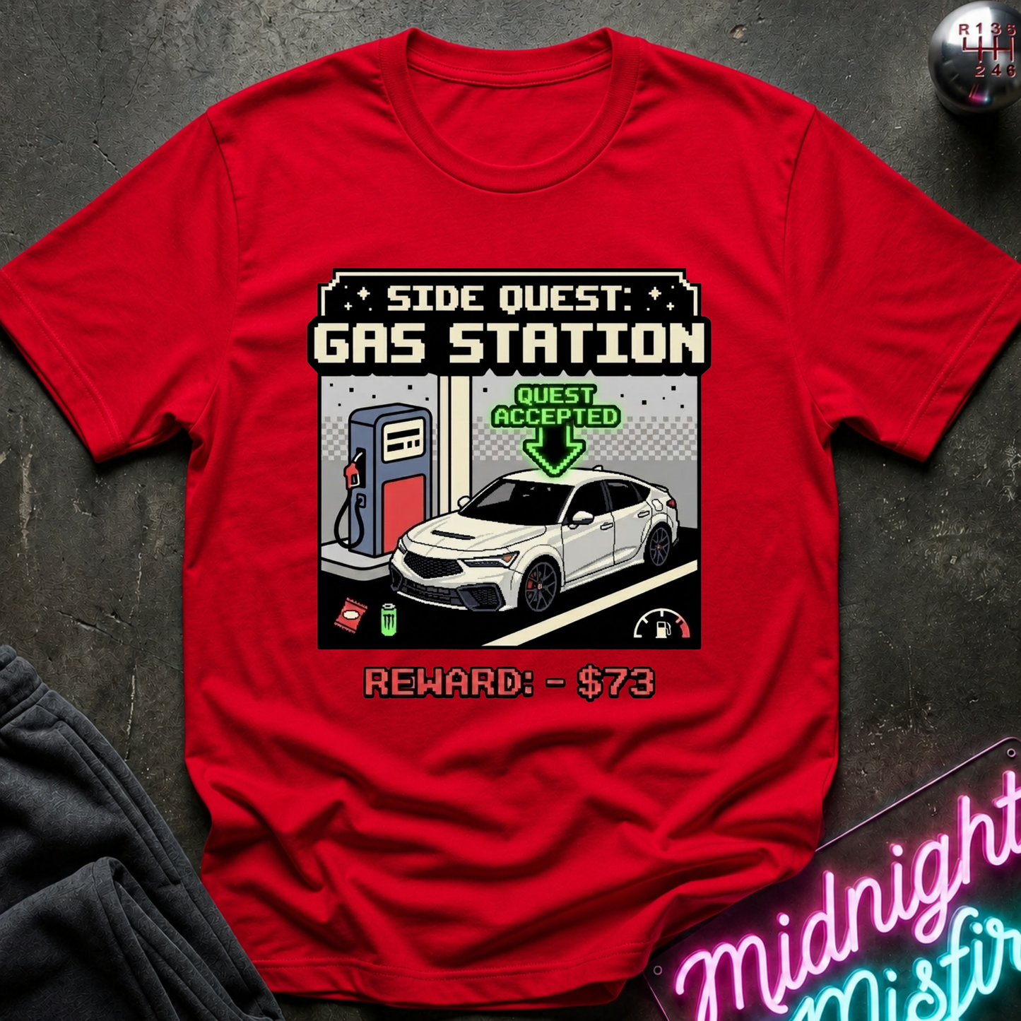 A red short-sleeve graphic T-shirt laid flat on a dark textured surface. The shirt features a pixel-art style gas station quest graphic with a white sedan, charging pump, and text reading 'SIDE QUEST GAS STATION' and 'REWARD - $73'.