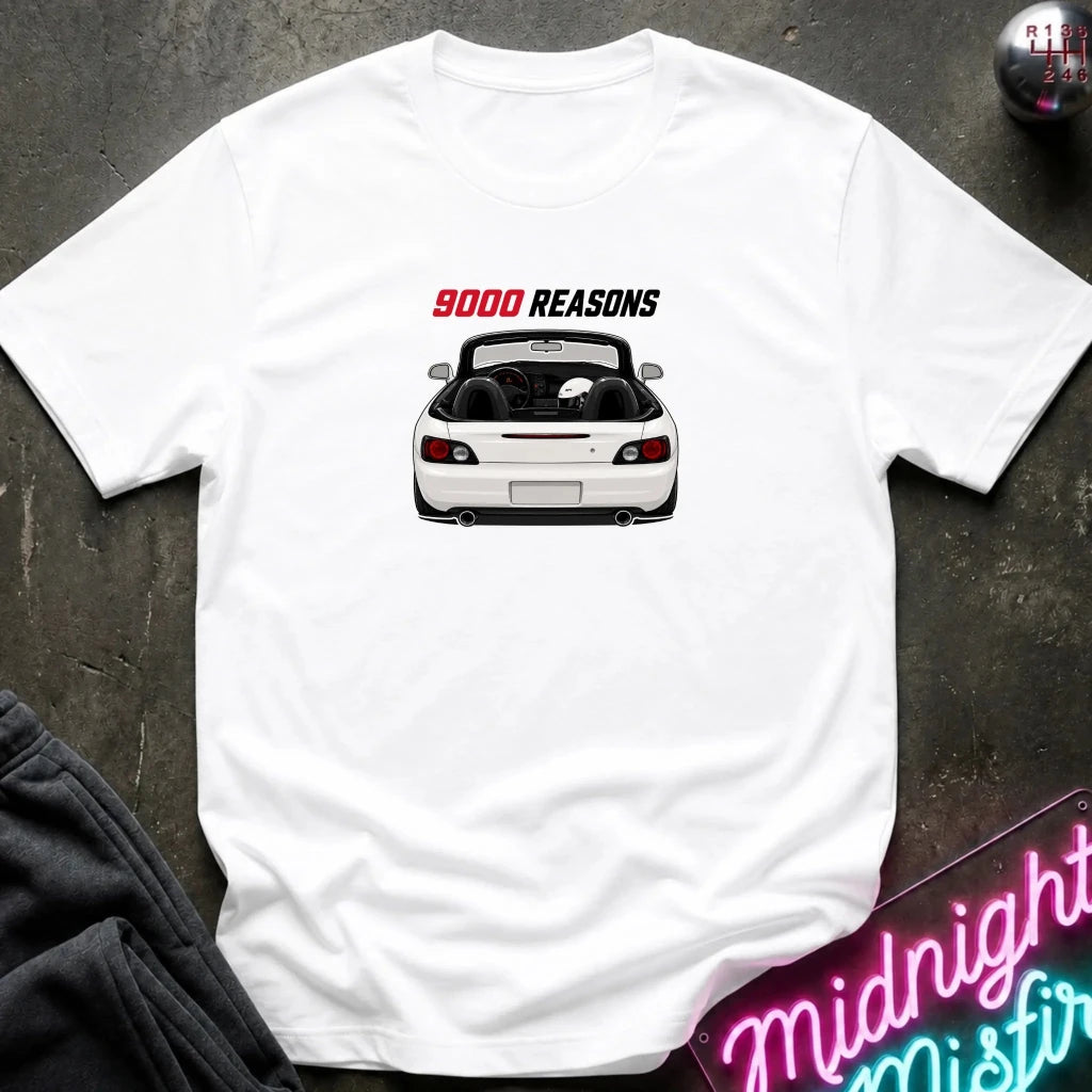 A white short-sleeve t-shirt laid flat on a dark textured surface, featuring a graphic of a white convertible sports car with the text '9000 REASONS' above it. A neon sign and dark clothing are partially visible in the background as styling props.