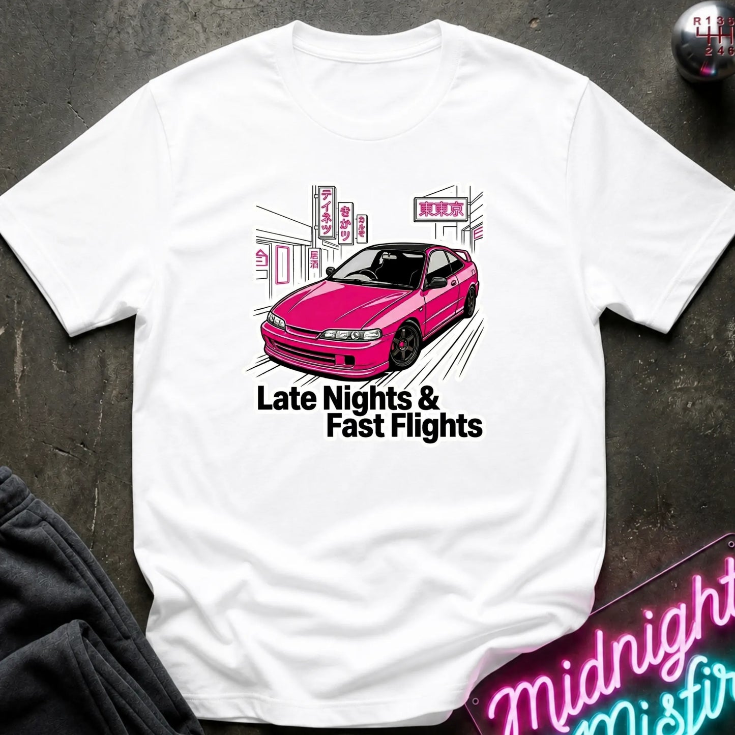 A white short-sleeve graphic T-shirt laid flat on a dark textured surface. The shirt features a pink car illustration with the text 'Late Nights & Fast Flights' printed below it.