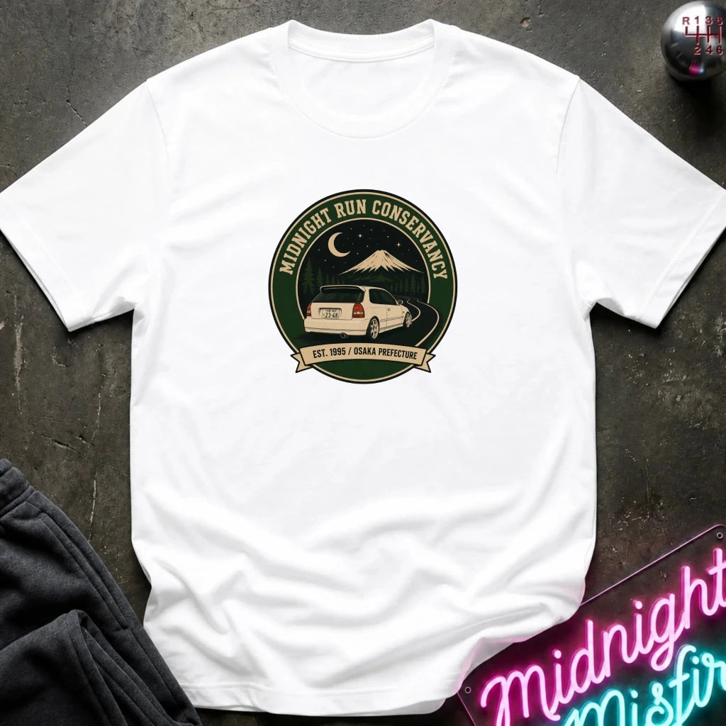 A white short-sleeve crewneck t-shirt laid flat on a dark textured surface. The center of the shirt features a circular graphic for 'Midnight Run Conservancy' with a mountain, moon, and a white SUV on a road, with the text 'EST.