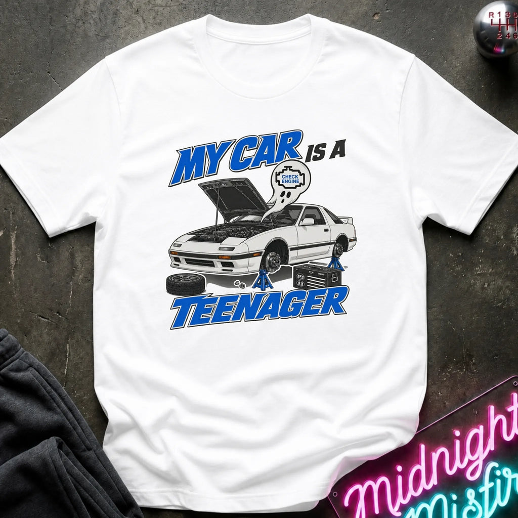 A white short-sleeve graphic T-shirt laid flat on a dark textured surface. The shirt features a cartoon-style car with its hood open, blue text reading 'MY CAR IS A TEENAGER,'.