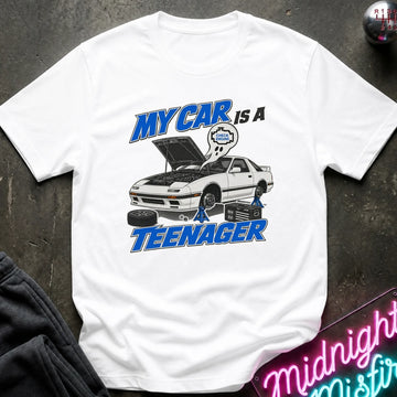 A white short-sleeve graphic T-shirt laid flat on a dark textured surface. The shirt features a cartoon-style car with its hood open, blue text reading 'MY CAR IS A TEENAGER,'.