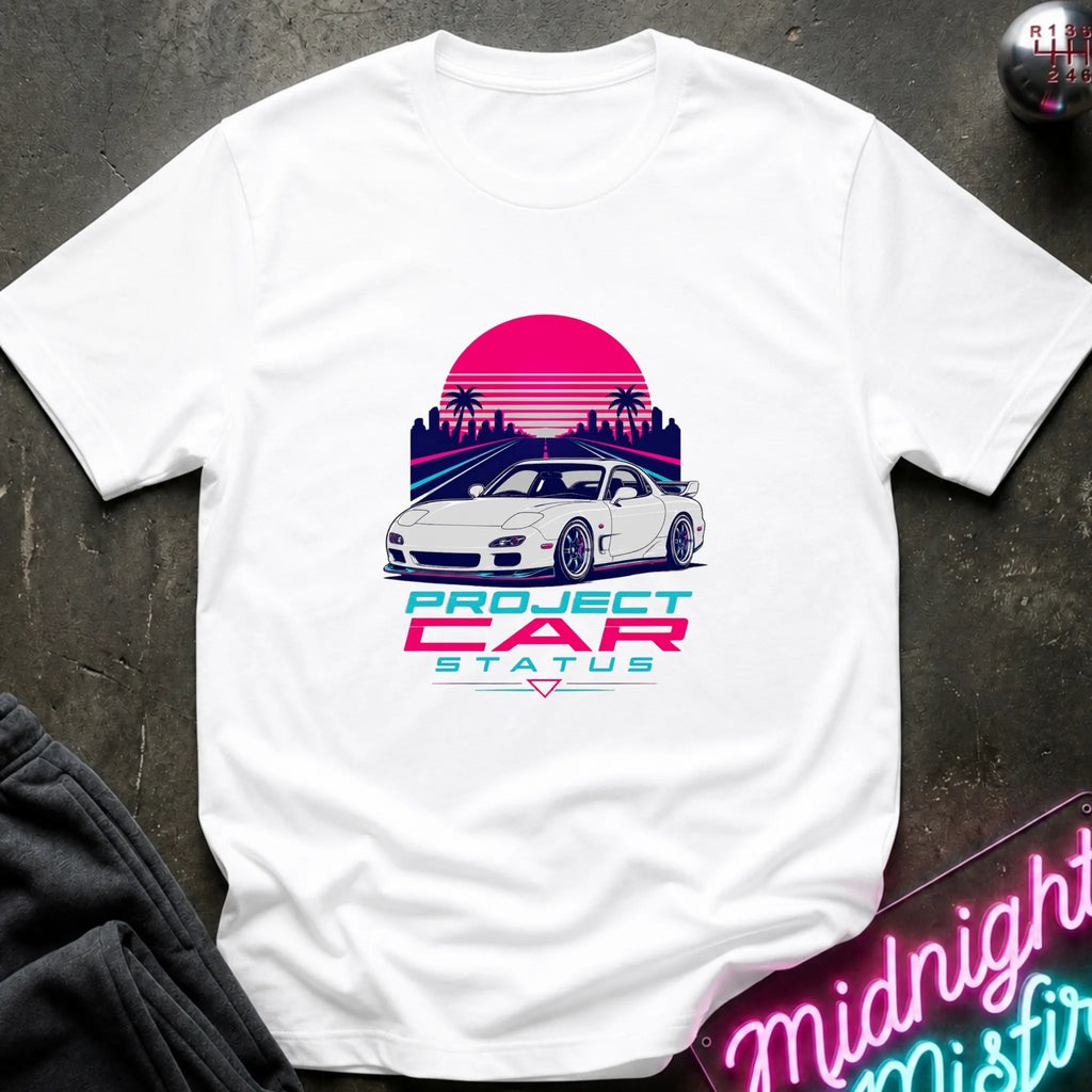 A white short-sleeve graphic T-shirt laid flat on a dark textured surface. The shirt features a retro-style print of a white sports car in front of a pink and purple sunset with palm trees, and the text 'PROJECT CAR STATUS' below.