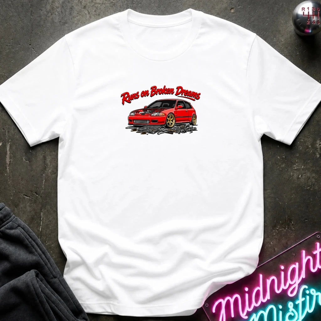 white crewneck t-shirt laid flat on a dark textured surface. The shirt features a graphic print of a red car with the text 'Run on Broken Dreams' in red and white script.