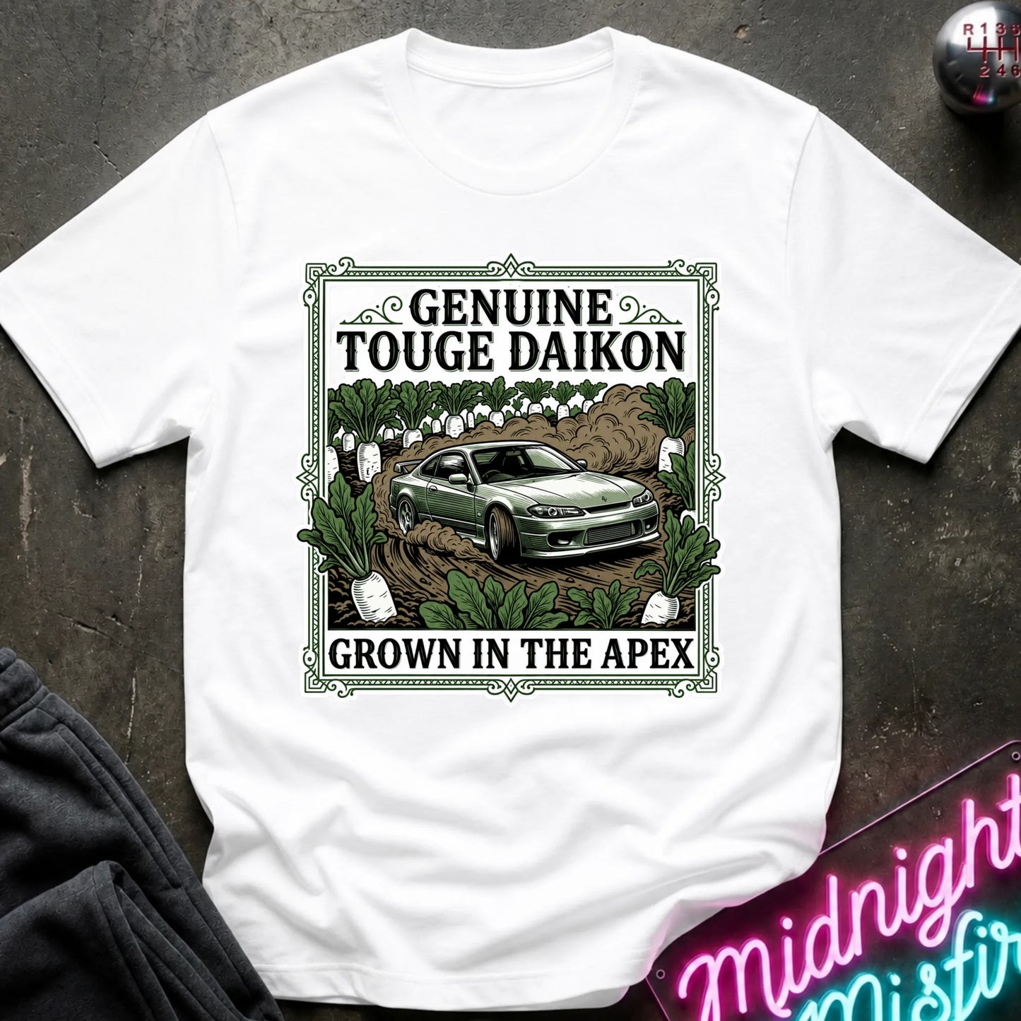 A white short-sleeve graphic T-shirt laid flat on a dark textured surface. The shirt features a large rectangular front print with the words 'GENUINE TOUGHE DAIKON' and 'GROWN IN THE APEX'.