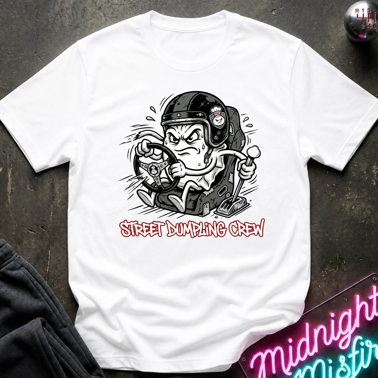 A white graphic t-shirt laid flat on a dark textured surface. The shirt features a cartoon illustration of a street-dumping character in a helmet with the text 'STREET DUMPING CREW' printed below in red and white.