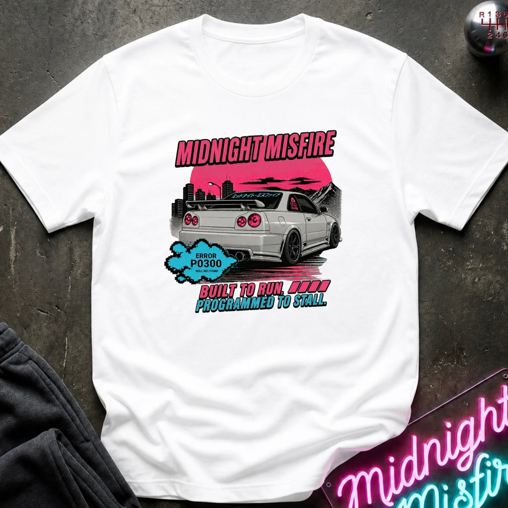 A white short-sleeve graphic t-shirt laid flat on a dark textured surface. The graphic features a white sports car with a pink sunset, the text 'MIDNIGHT MISFIRE' in pink.