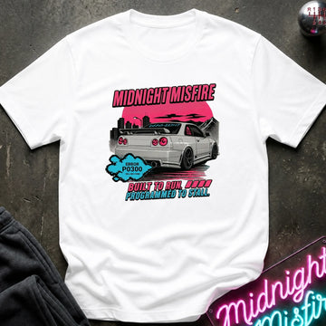 A white short-sleeve graphic t-shirt laid flat on a dark textured surface. The graphic features a white sports car with a pink sunset, the text 'MIDNIGHT MISFIRE' in pink.