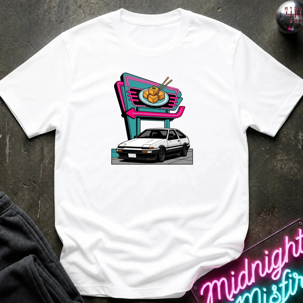 A white short-sleeve t-shirt laid flat on a dark textured surface, featuring a colorful graphic print of a white hatchback parked in front of a neon-style food sign. 