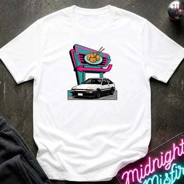 A white short-sleeve t-shirt laid flat on a dark textured surface, featuring a colorful graphic print of a white hatchback parked in front of a neon-style food sign. 