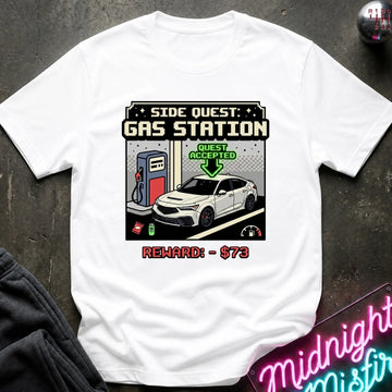 A white  graphic t-shirt laid flat on a dark textured surface. The shirt features a retro-futuristic pixel-art style print of a gas station with a white sports car, a fuel pump, and text reading 'SIDE QUEST: GAS STATION', 'QUEST ACCEPTED', and 'REWARD: - $73'.