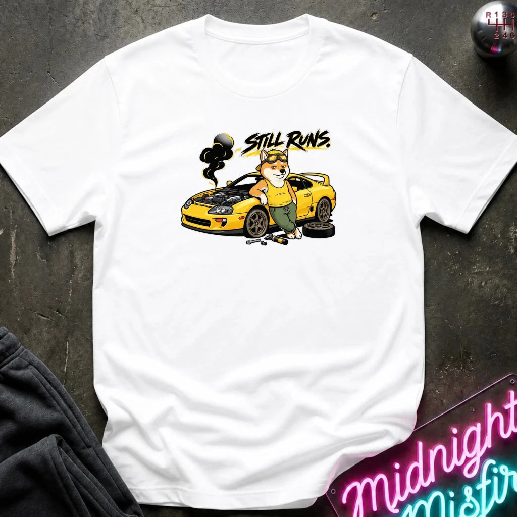 white short-sleeve graphic t-shirt laid flat on a dark textured surface. The graphic features a cartoon dog in a mechanic hat standing next to a yellow sports car with the text 'Still runs' above it.