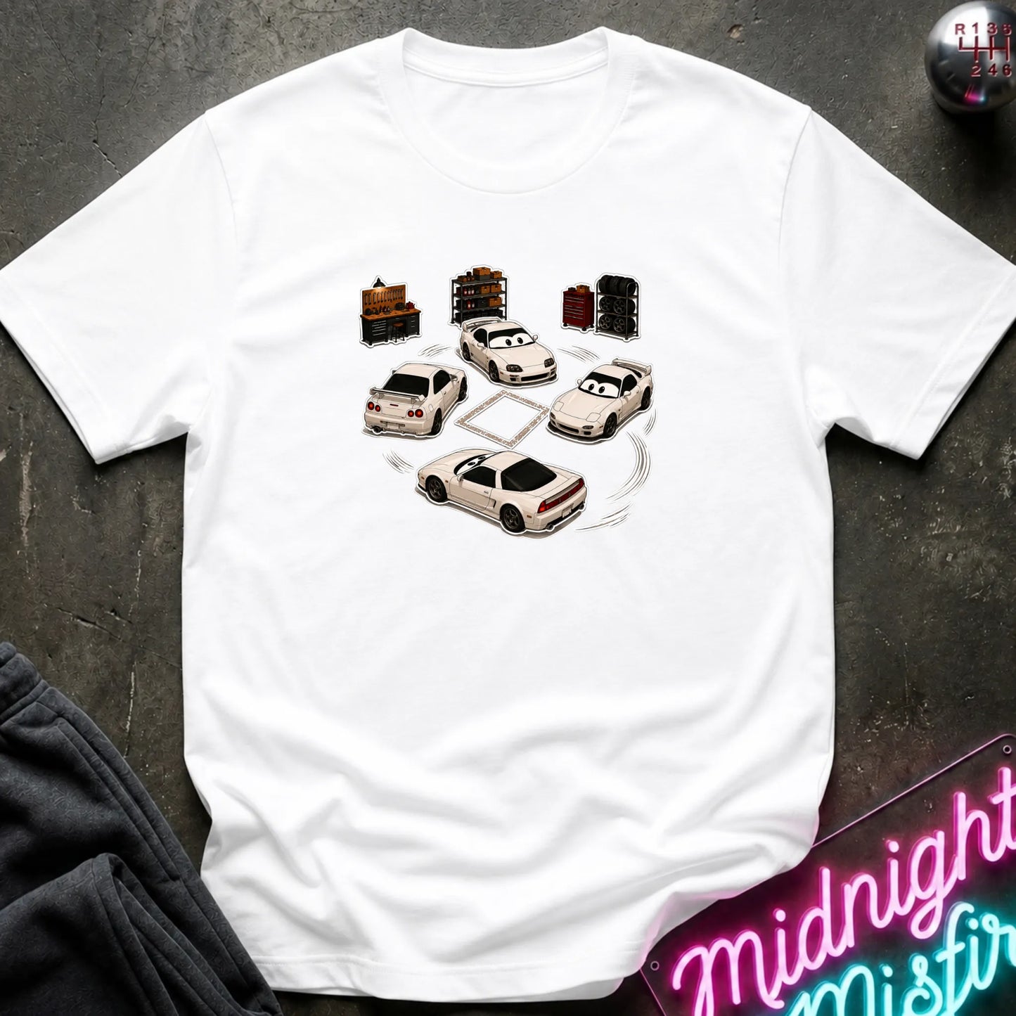 A white short-sleeve t-shirt laid flat on a dark textured surface, featuring a graphic print of several white sports cars arranged in a circle with drift-like tire marks.