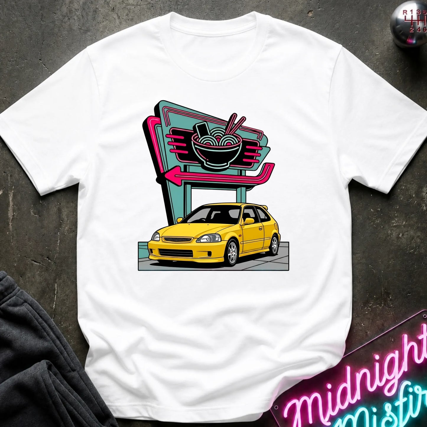 A white short-sleeve crewneck t-shirt laid flat on a dark textured surface, featuring a colorful graphic print of a yellow car parked in front of a retro neon ramen shop sign.
