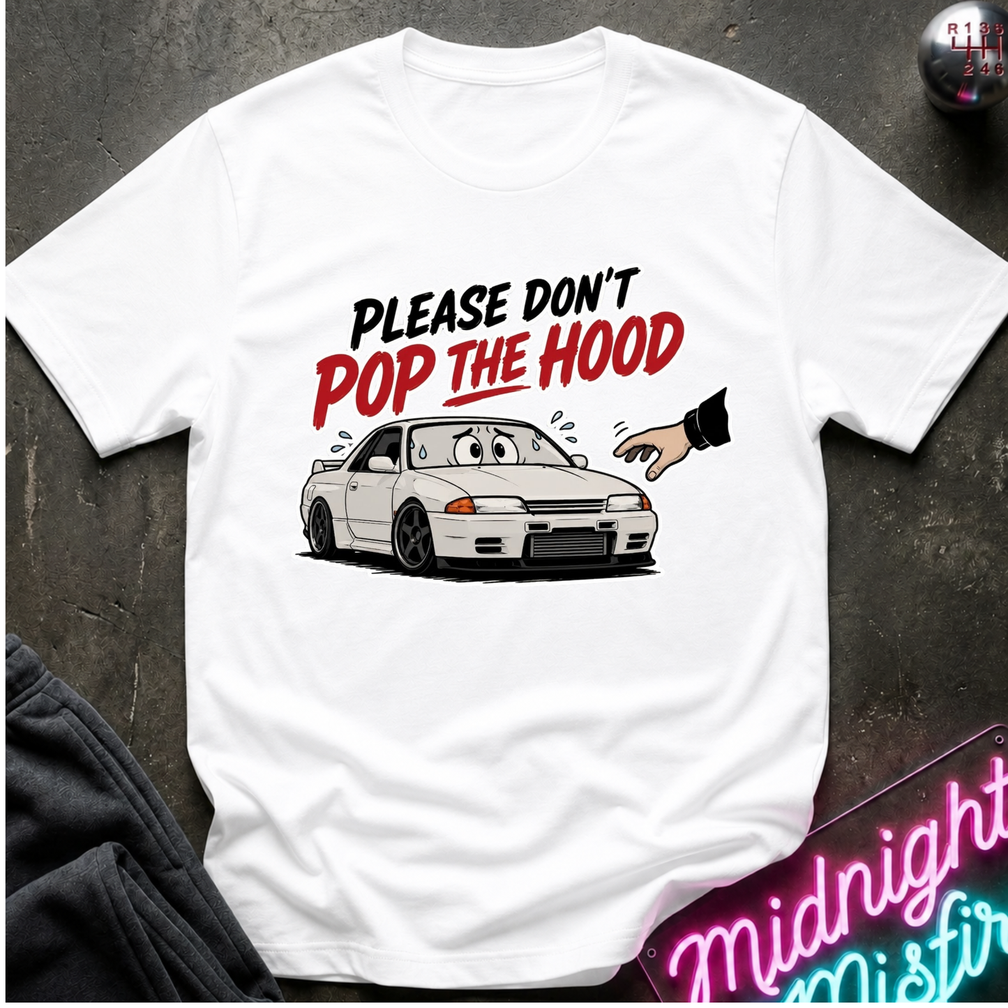 A white short-sleeve graphic T-shirt laid flat on a dark textured surface. The shirt features a cartoon illustration of a white sports car with a surprised face, a hand pointing at it, and the text 'PLEASE DON'T POP THE HOOD' in black and red lettering.