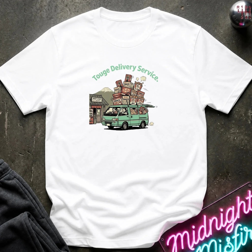 A white short-sleeve t-shirt laid flat on a dark textured surface, featuring a colorful graphic of a green van carrying stacked boxes with the text 'Touge Delivery Service.' printed above it.