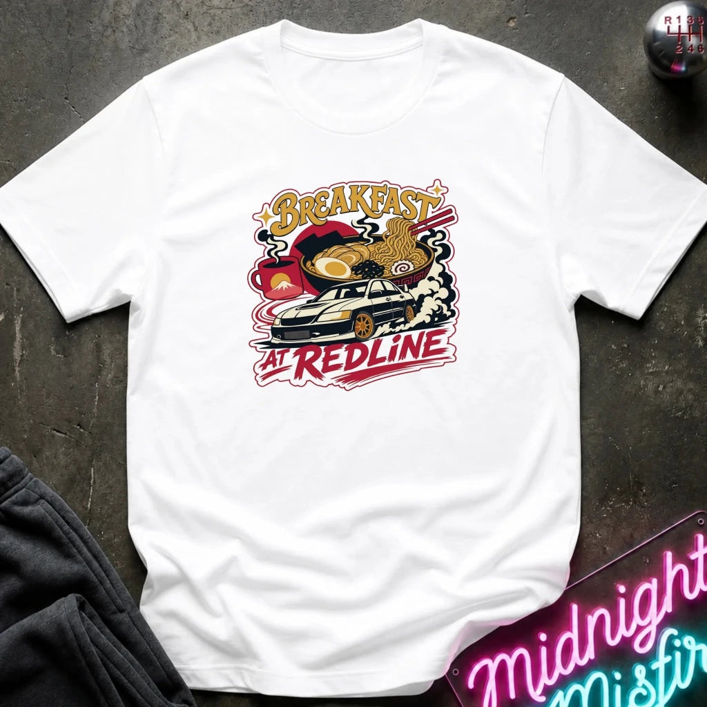 A white short-sleeve graphic T-shirt laid flat on a dark textured surface. The shirt features a colorful retro-style chest print with the words 'Breakfast at Redline'