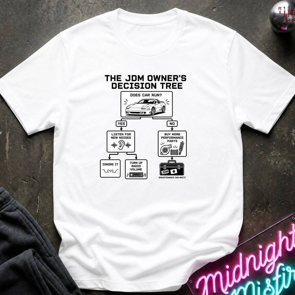A white short-sleeve graphic T-shirt laid flat on a dark textured surface. The shirt features a black printed decision tree titled 'THE JDM OWNER'S DECISION TREE' with illustrations and text related to car care and maintenance.