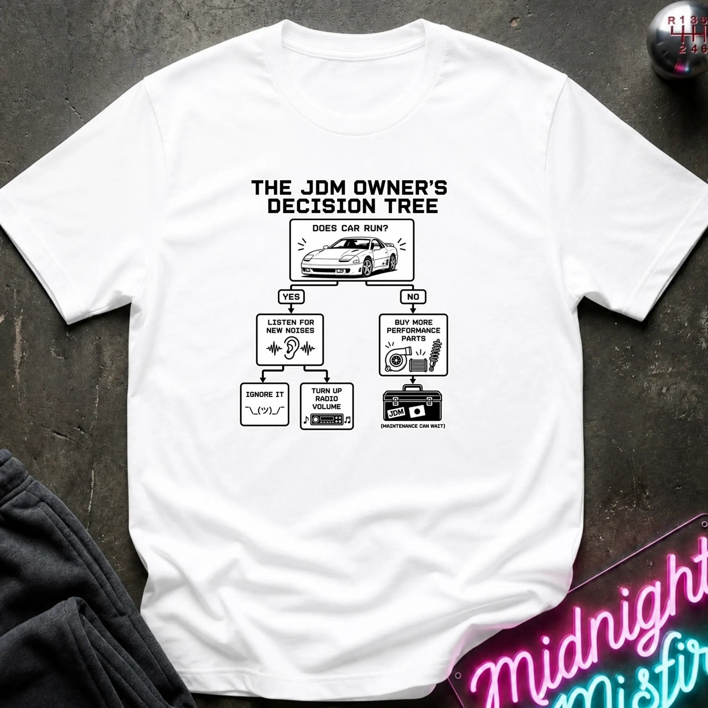 A white short-sleeve graphic T-shirt laid flat on a dark textured surface. The shirt features a black printed decision tree titled 'THE JDM OWNER'S DECISION TREE' with illustrations and text related to car care and maintenance.