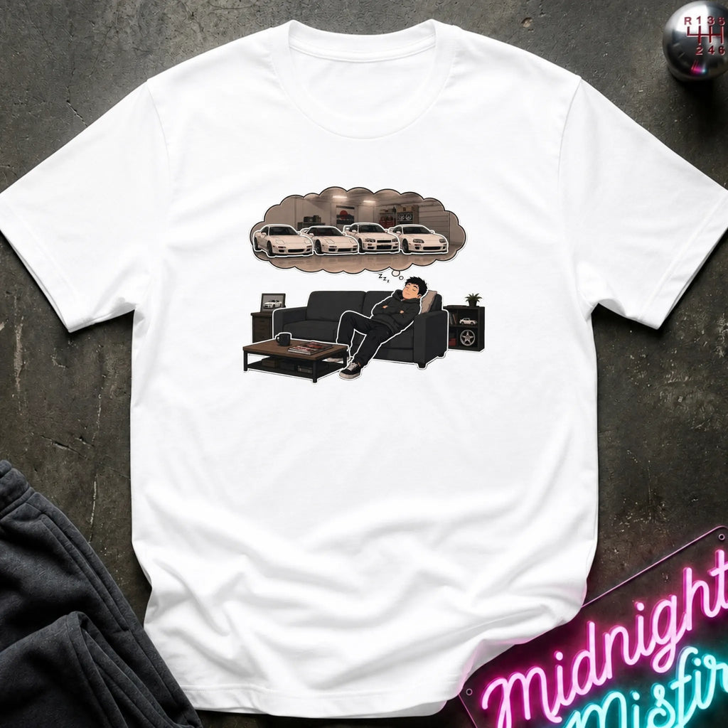 A white short-sleeve graphic t-shirt laid flat on a dark textured surface. The graphic shows a man lounging on a black couch with a thought bubble above his head containing white sports cars.