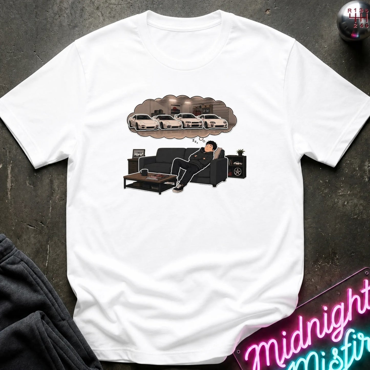 A white short-sleeve graphic t-shirt laid flat on a dark textured surface. The graphic shows a man lounging on a black couch with a thought bubble above his head containing white sports cars.