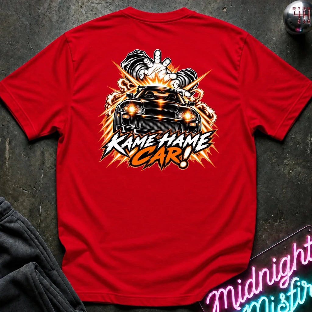 A red short-sleeve graphic T-shirt laid flat on a dark textured surface. The back of the shirt features a large graphic print of a black car with glowing orange headlights, flames, smoke, and the text 'KAME HAME CAR!' in stylized white and orange lettering.