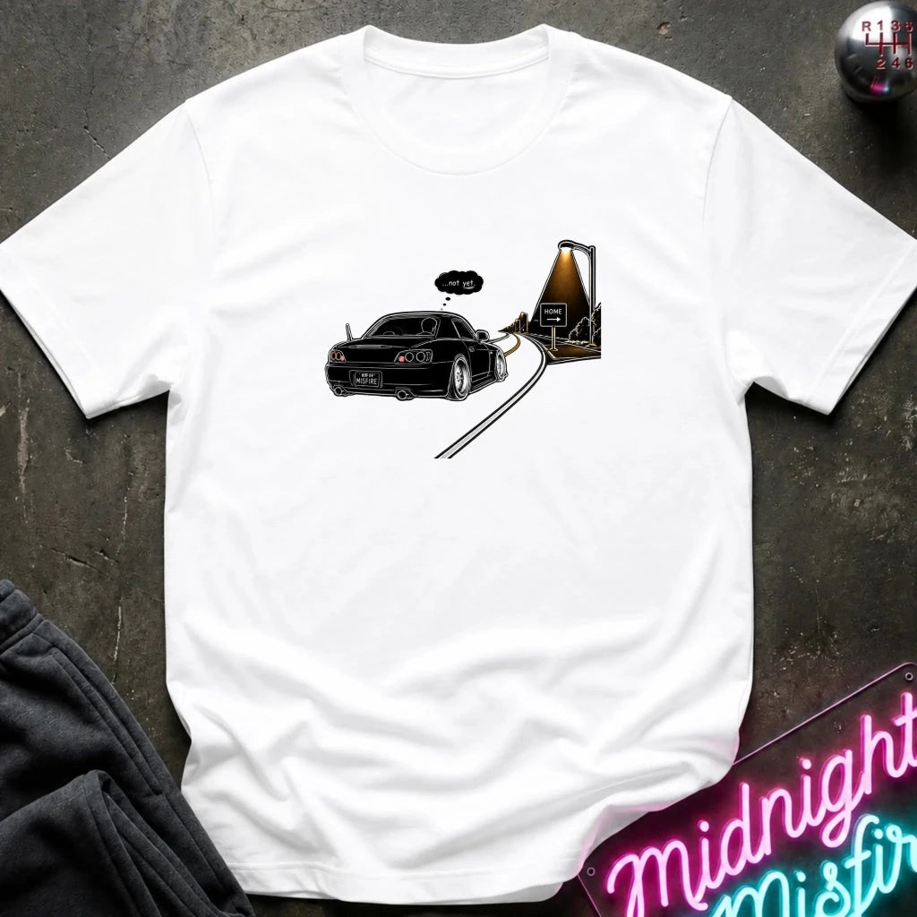 A white short-sleeve t-shirt laid flat on a dark textured surface, featuring a graphic print of a black sports car driving passed sign that says home.