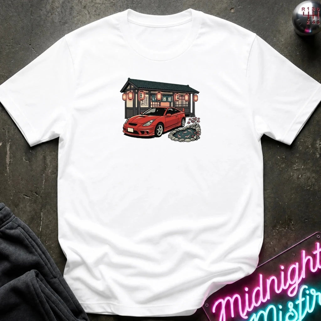 A white short-sleeve t-shirt laid flat on a dark textured surface, featuring a graphic print of a red sports car parked in front of a Japanese-style building. 
