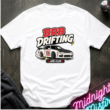 A white short-sleeve graphic T-shirt laid flat on a dark textured surface. The shirt features a large 'BRB DRIFTING' print in red, black, and white above an illustration of a drift car with 'JDM CLUB' text.