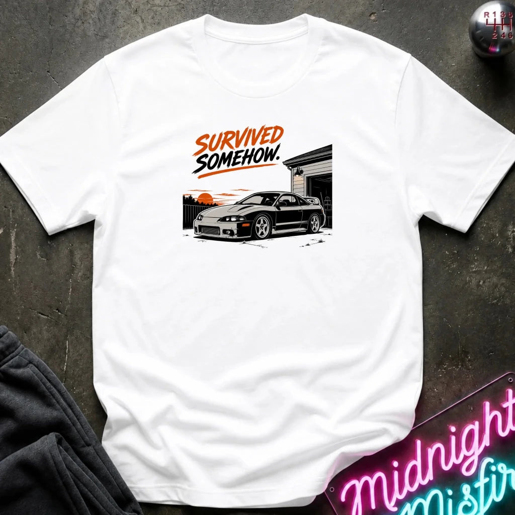 A white short-sleeve graphic T-shirt laid flat on a dark textured surface. The shirt features a printed design of a black sports car in front of a garage with orange and black text reading 'SURVIVED SOMEHOW.