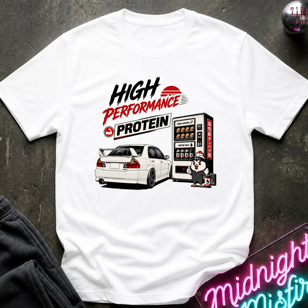 A white short-sleeve graphic T-shirt laid flat on a dark textured surface. The shirt features a printed design with the words 'HIGH PERFORMANCE PROTEIN' in black and red, an illustration of a white sports car next to a vending machine, and a small character.