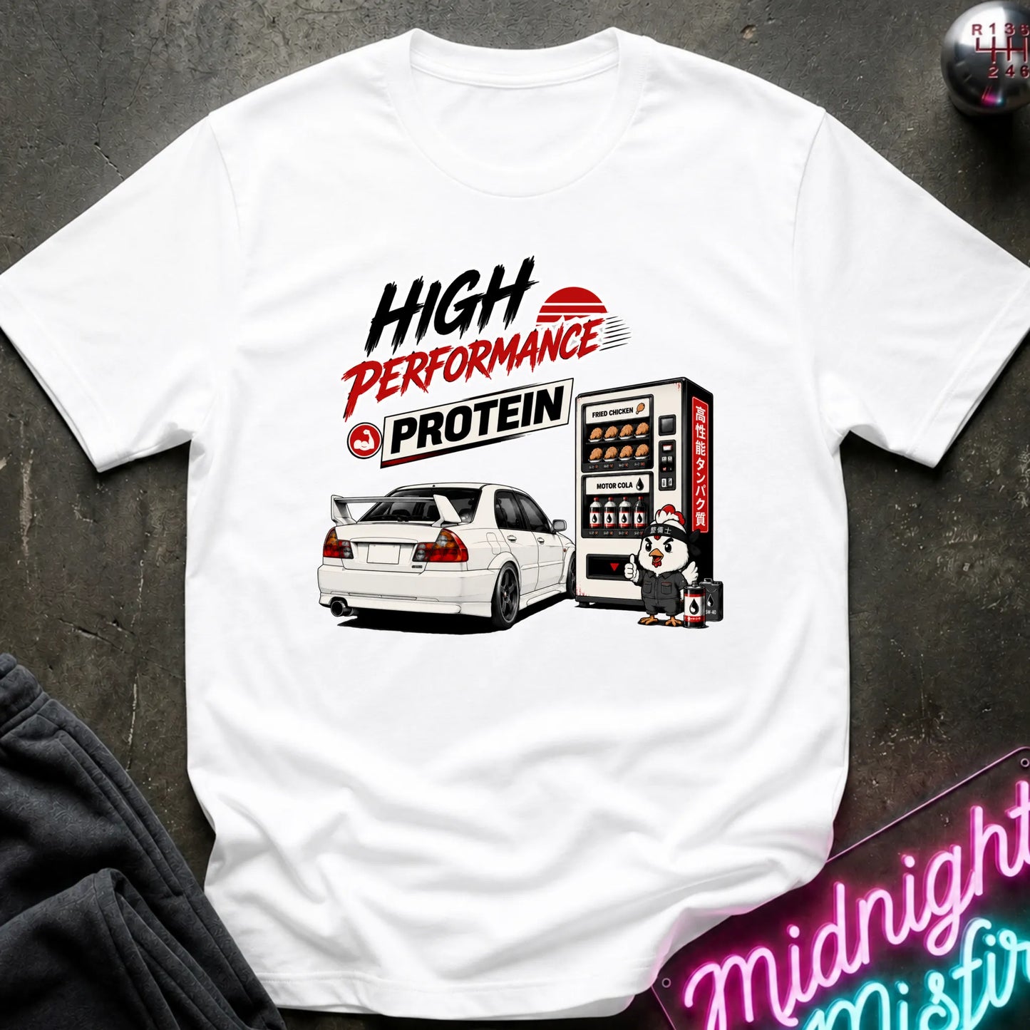 A white short-sleeve graphic T-shirt laid flat on a dark textured surface. The shirt features a printed design with the words 'HIGH PERFORMANCE PROTEIN' in black and red, an illustration of a white sports car next to a vending machine, and a small character.