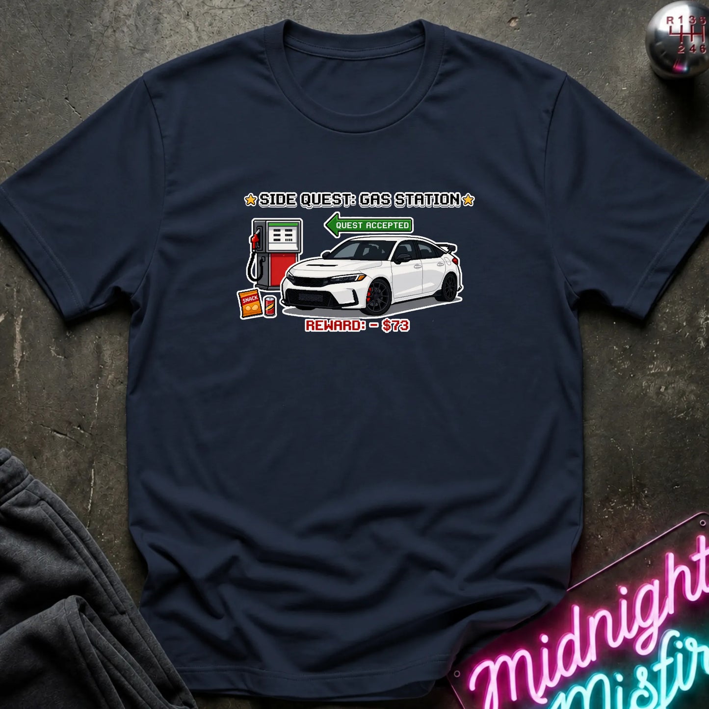 A navy blue short-sleeve t-shirt with a graphic print of a white sports car at a gas station, laid flat on a dark textured surface with a neon sign and folded jeans nearby.