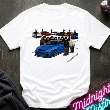 A white short-sleeve t-shirt laid flat on a dark textured surface, featuring a colorful graphic print of a blue sports car and a group of people in the center, with smaller cars in the background. A neon sign and dark clothing are partially visible around the edges.