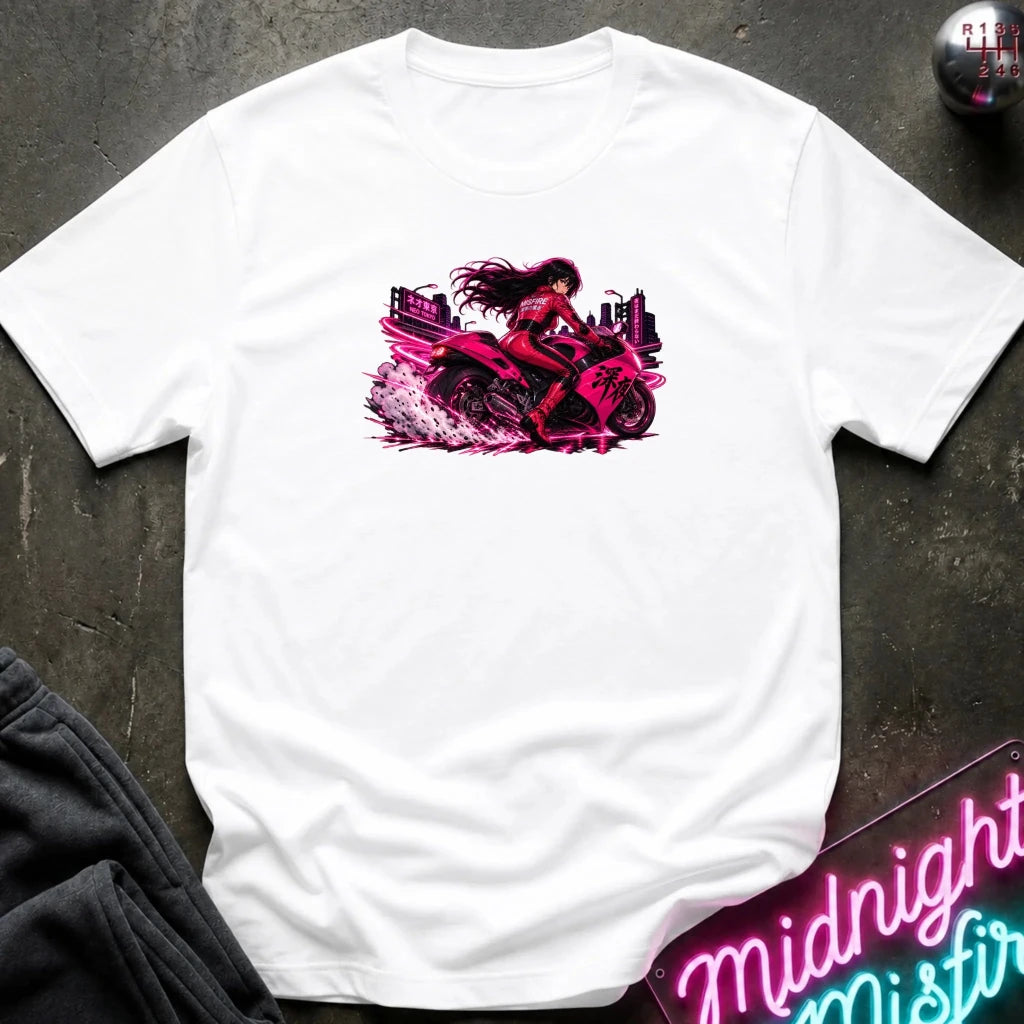 A white short-sleeve t-shirt laid flat on a dark textured surface, featuring a vibrant graphic print of a woman riding a pink motorcycle. The shirt is styled with a pair of black pants in the lower-left corner and a neon sign in the lower-right corner.