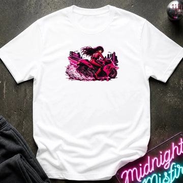 A white short-sleeve t-shirt laid flat on a dark textured surface, featuring a vibrant graphic print of a woman riding a pink motorcycle. The shirt is styled with a pair of black pants in the lower-left corner and a neon sign in the lower-right corner.