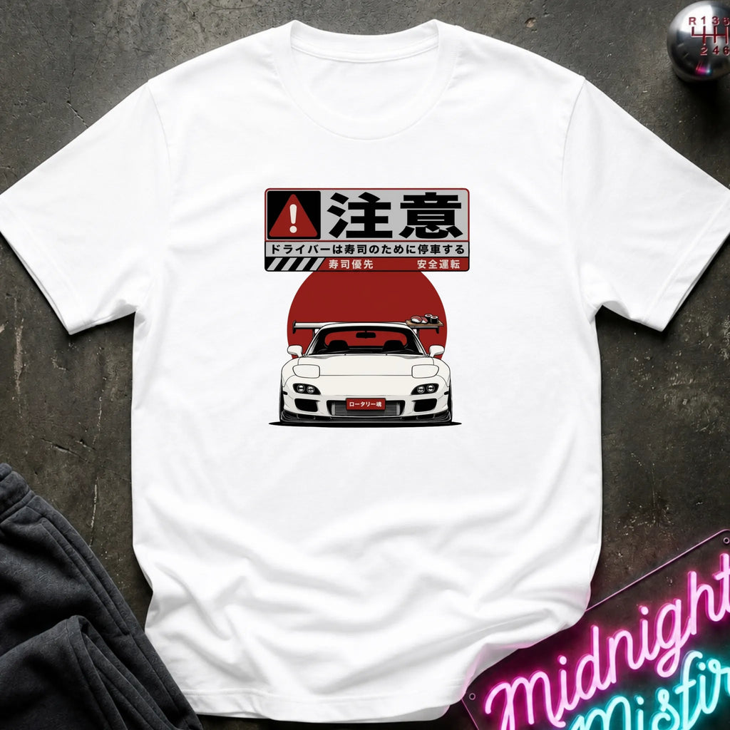 A white short-sleeve graphic t-shirt laid flat on a dark textured surface. The graphic features a white sports car in front of a large red sun with Japanese text above it.