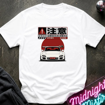A white short-sleeve graphic t-shirt laid flat on a dark textured surface. The graphic features a white sports car in front of a large red sun with Japanese text above it.