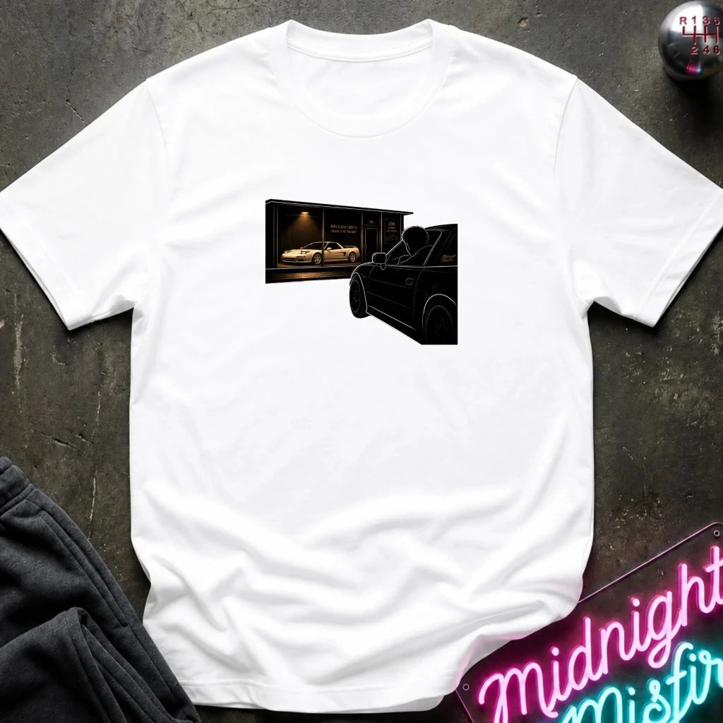A white short-sleeve t-shirt laid flat on a dark textured surface, featuring a rectangular graphic print on the chest of a black car silhouette in the foreground and a white sports car inside a garage in the background.