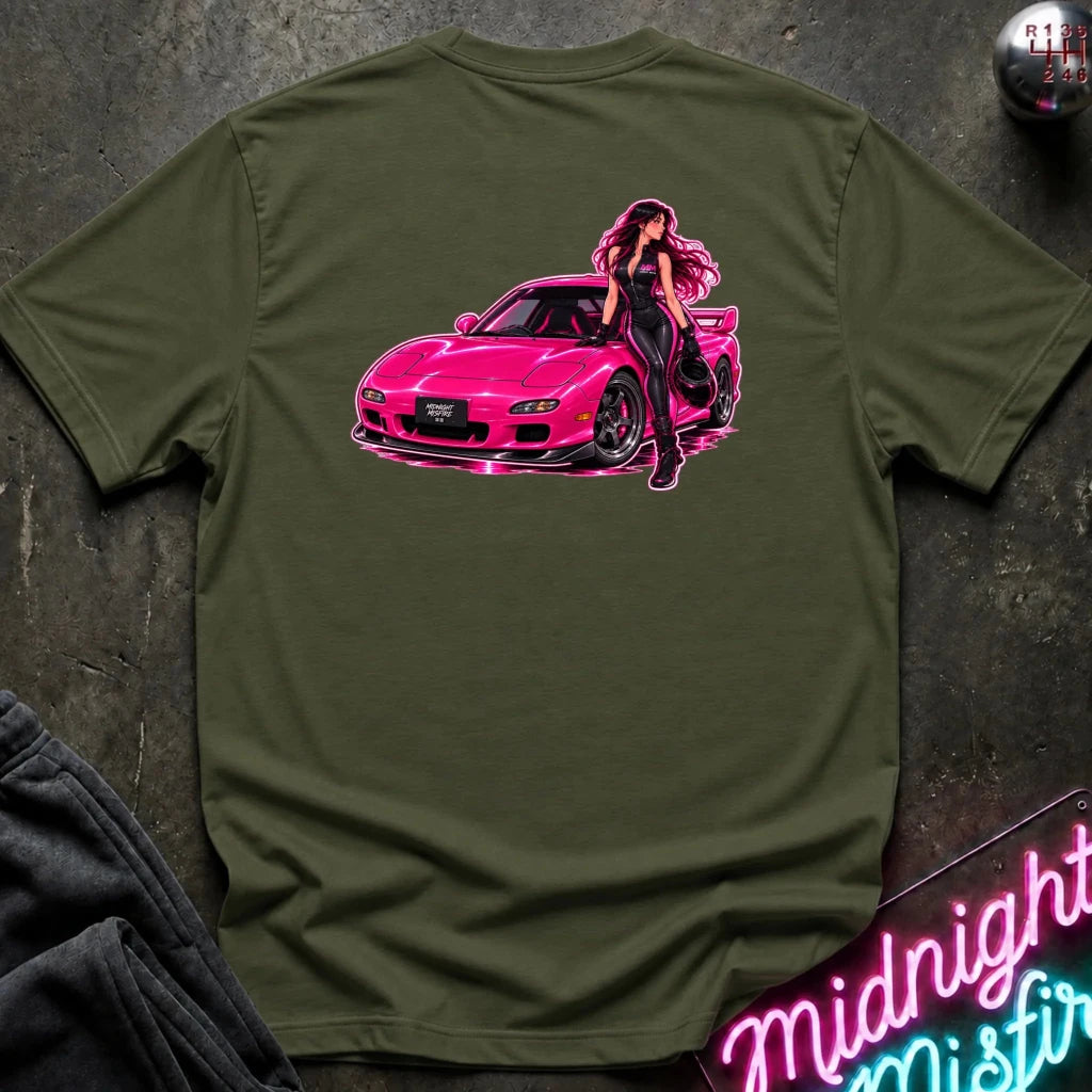 An olive green short-sleeve t-shirt laid flat on a dark textured surface, featuring a graphic print of a woman with pink hair leaning against a bright pink sports car. The shirt is styled with a dark gray sweatshirt in the corner and a neon sign in the background.