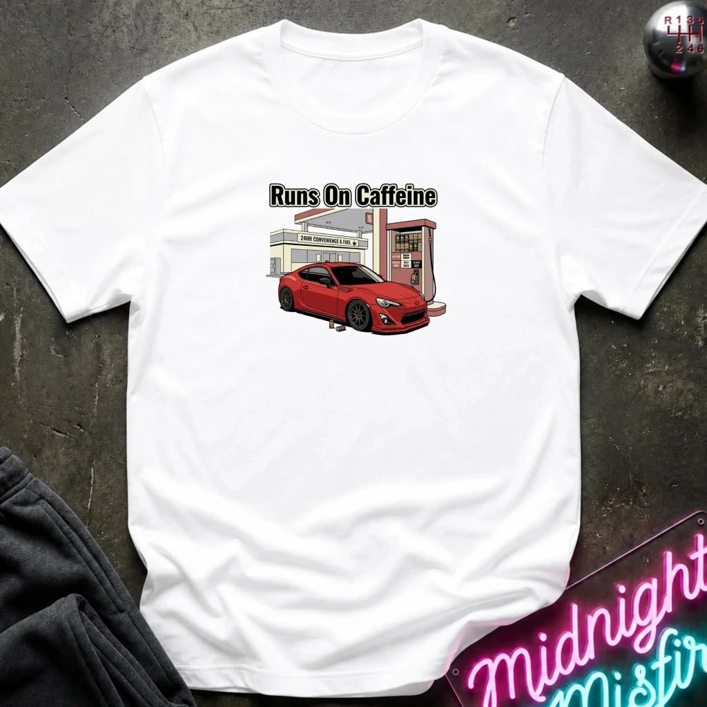 A white short-sleeve t-shirt laid flat on a dark textured surface, featuring a graphic print of a red sports car at a gas station with the text 'Runs On Caffeine'.