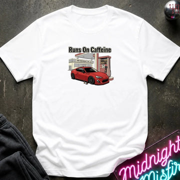 A white short-sleeve t-shirt laid flat on a dark textured surface, featuring a graphic print of a red sports car at a gas station with the text 'Runs On Caffeine'.